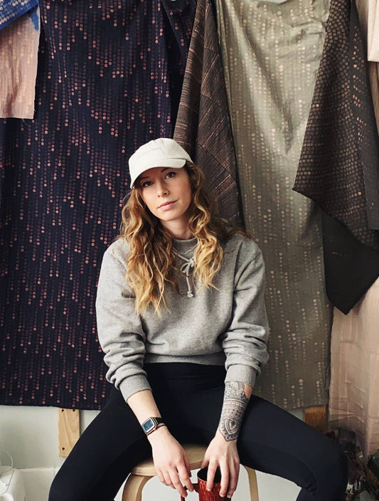 Cassie sits in her studio, wearing a hat and holding a mug. 